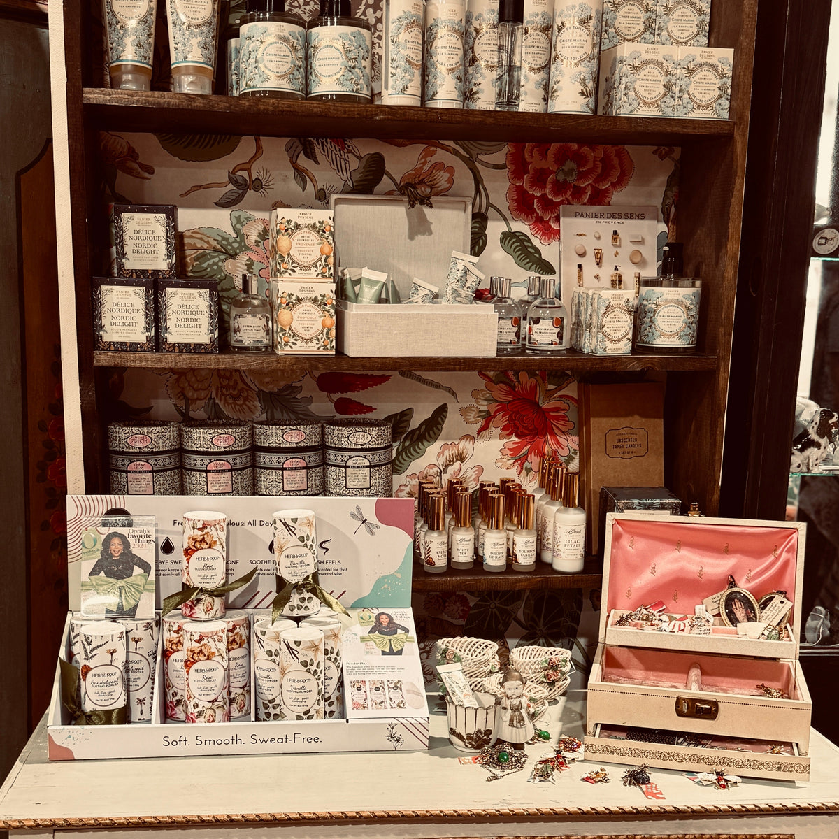 A styled display of various bath and body products, candles, and accessories arranged on a white vintage-style cabinet against a floral wallpapered background. The collection includes patterned toiletries, candles in a pink-lidded box, and small bottles.