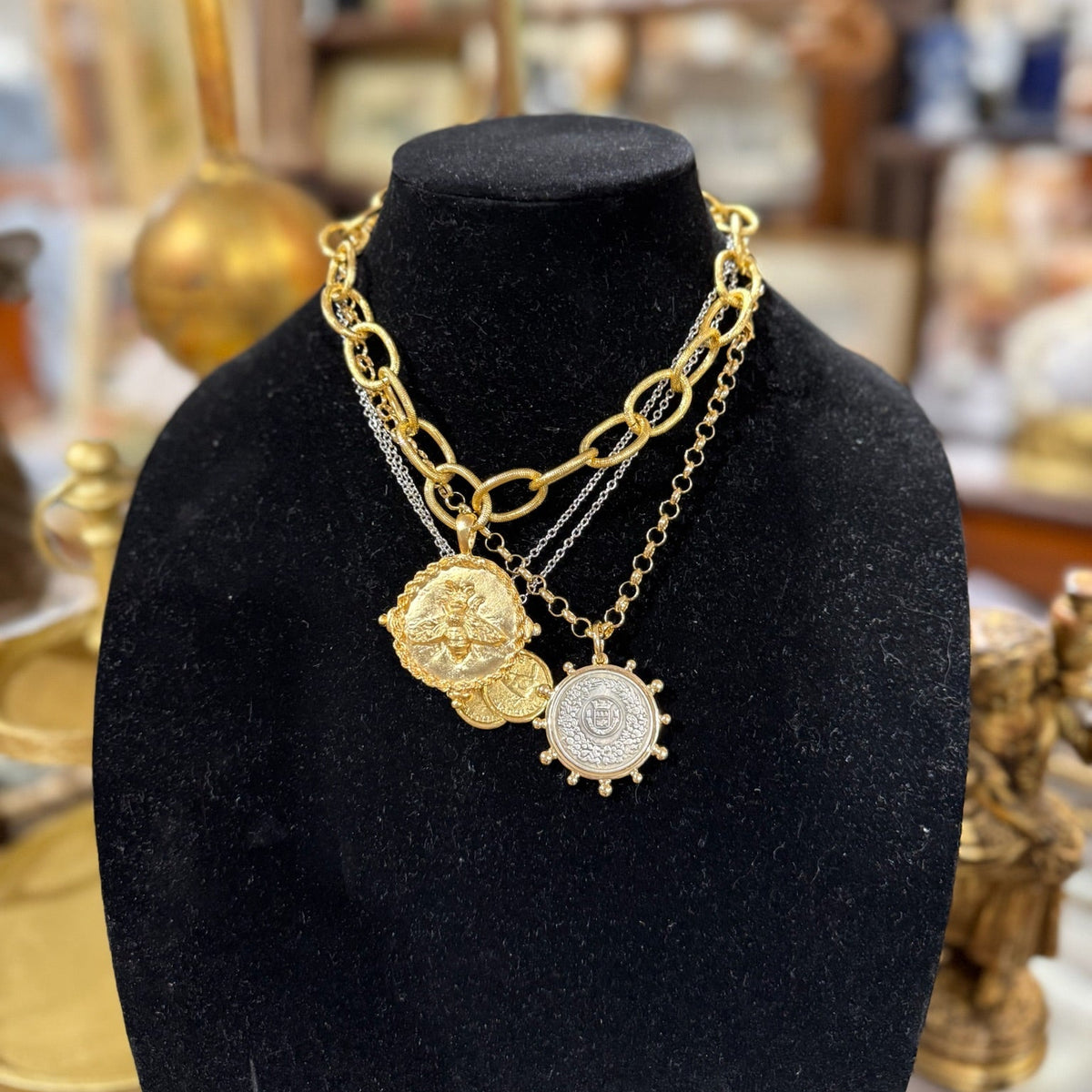 A layered gold and silver chain necklace with three distinct pendants, displayed on a black velvet jewelry bust. The necklace features a chunky gold chain, a delicate silver chain, a gold coin pendant, and a gold ship's wheel pendant.