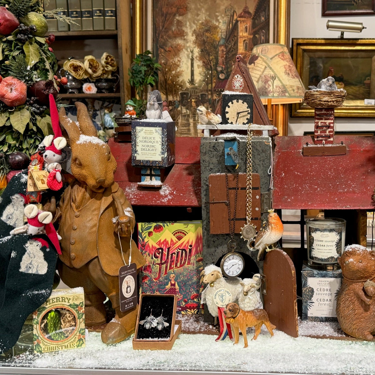A dense, festive tabletop display filled with holiday decorations, figurines, vintage-style items, and ornaments, including a rabbit statue, Christmas trees, a small clock tower, and assorted decorative objects arranged on a snowy-white surface.